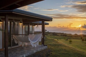 A hammock under a covered porch with a view of the sunset over a grassy landscape.