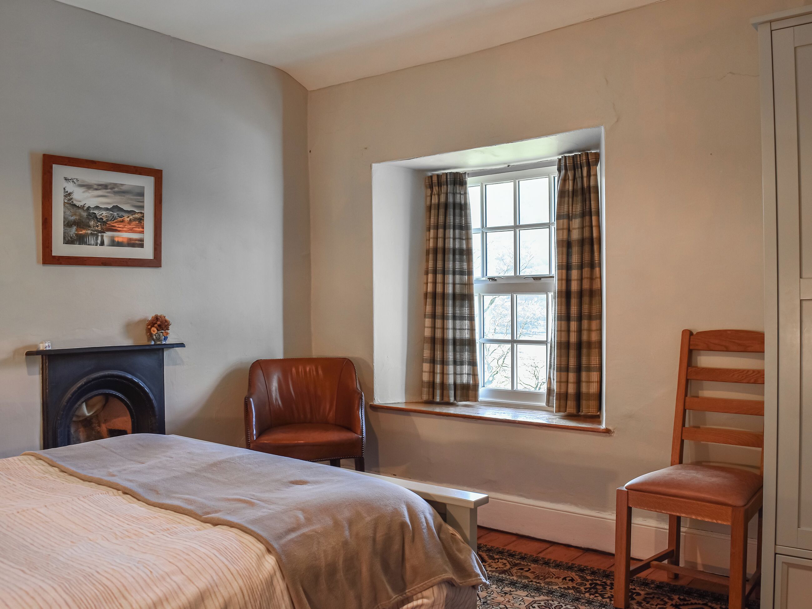 A room with a bed, a fireplace, a window with curtains, a chair, and a wooden chair.