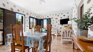 A dining area with a table set for four, surrounded by wooden chairs, a television mounted on the wall, and a potted plant.