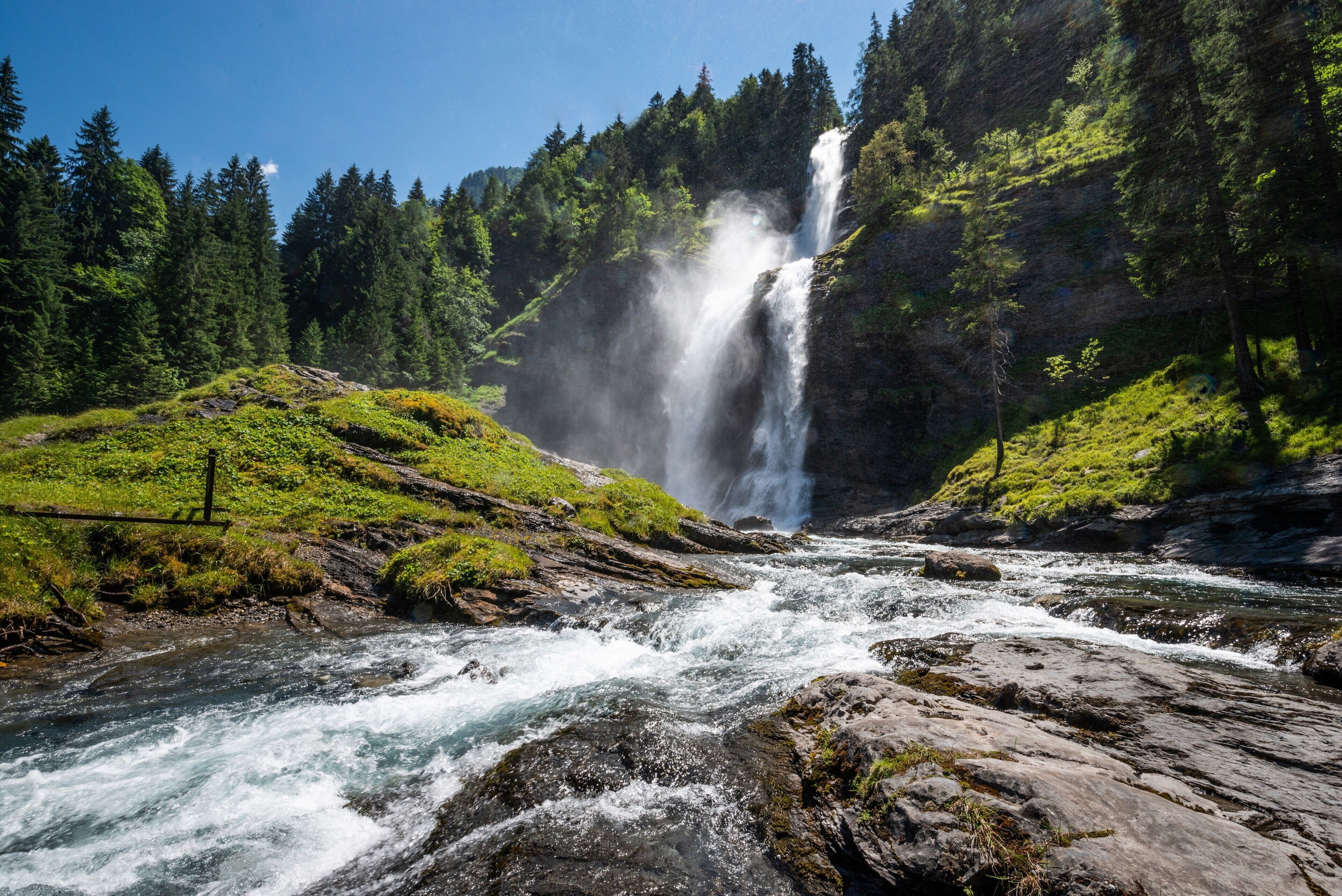 Ein Wasserfall stürzt in einen Fluss, umgeben von bewaldeten Hügeln.