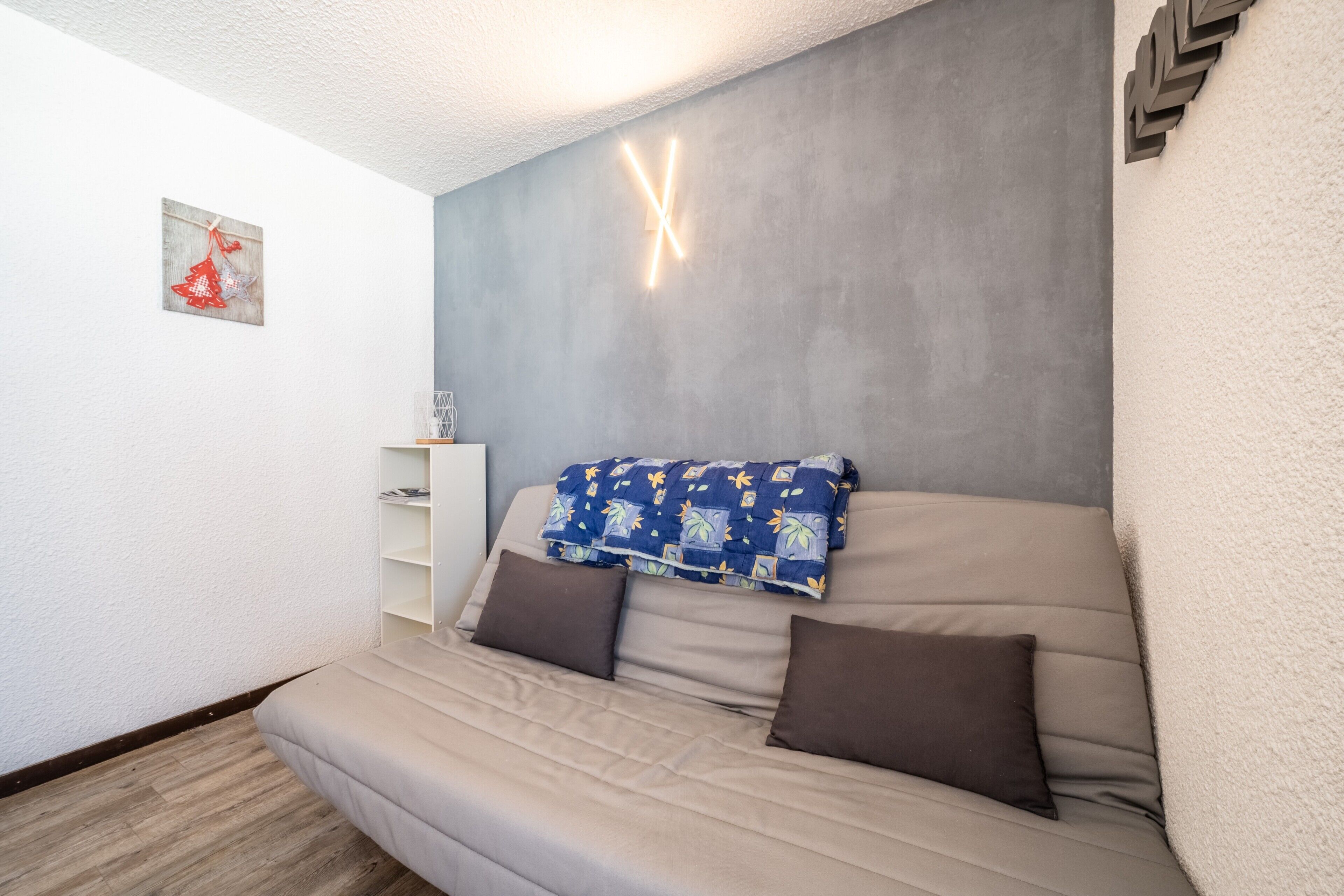Een kamer met een slaapbank, een klein wit schap en een wandlamp.