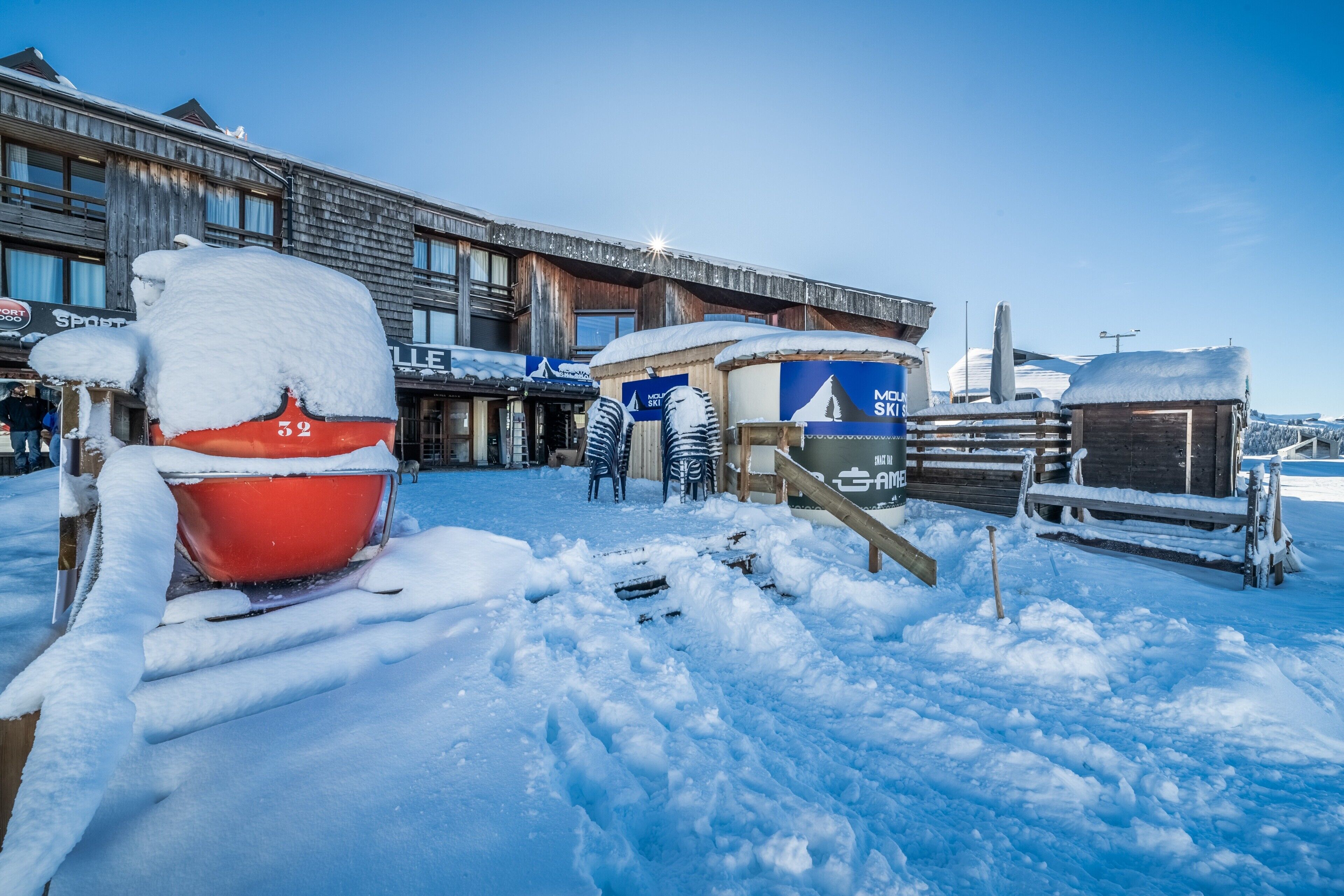 Un espace extérieur enneigé avec un baril rouge, un bâtiment bleu portant une enseigne et une structure en bois.