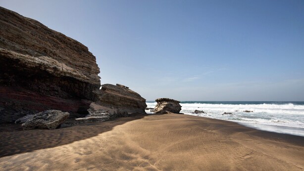Ein Strand mit einer felsigen Klippe, Wellen und blauem Himmel.
