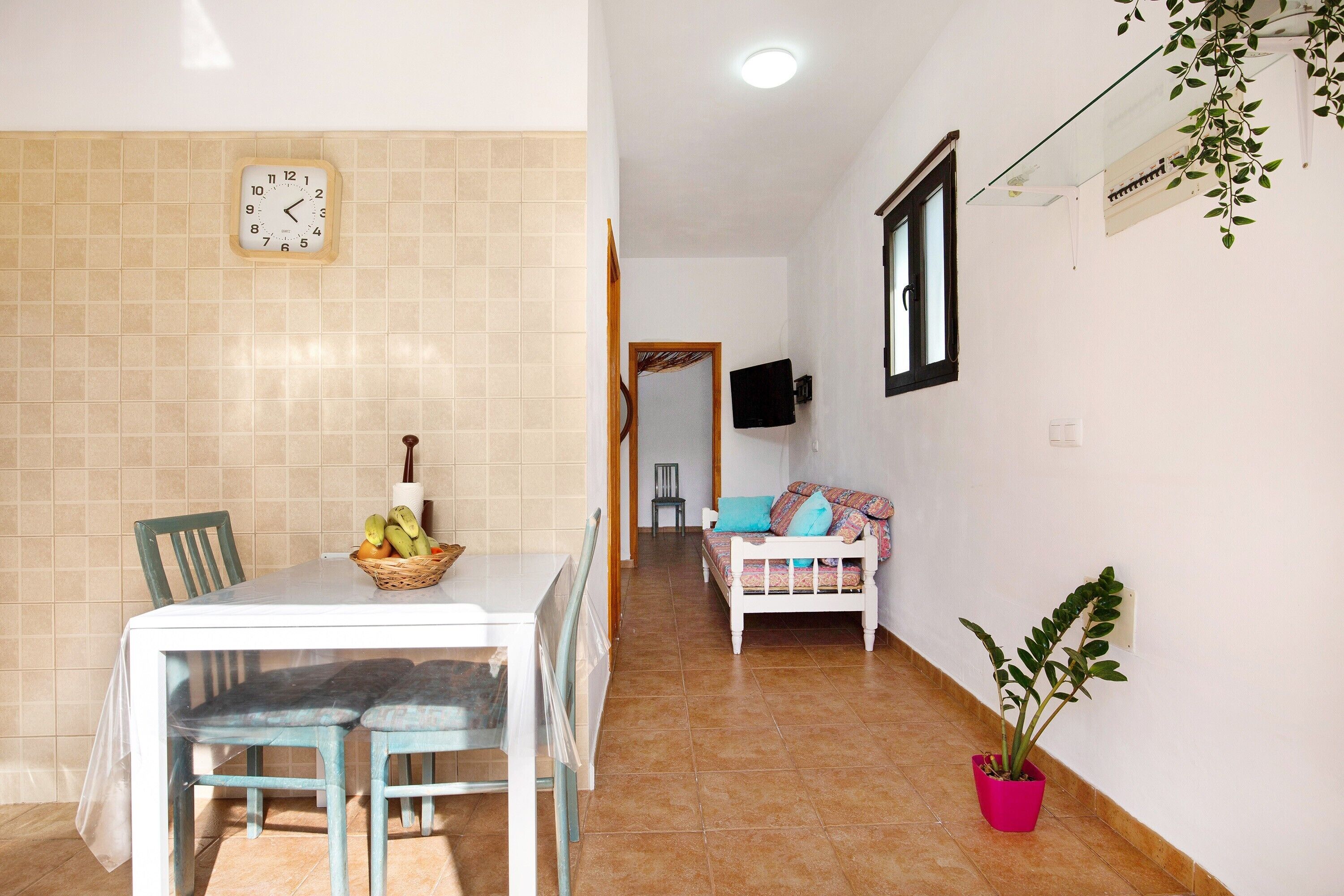 A hallway with a dining table, chairs, a sofa, a potted plant, and a wall clock.