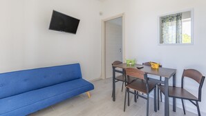 Una habitación con un sofá azul, una mesa de comedor con sillas, un televisor montado en la pared y una ventana con cortinas.