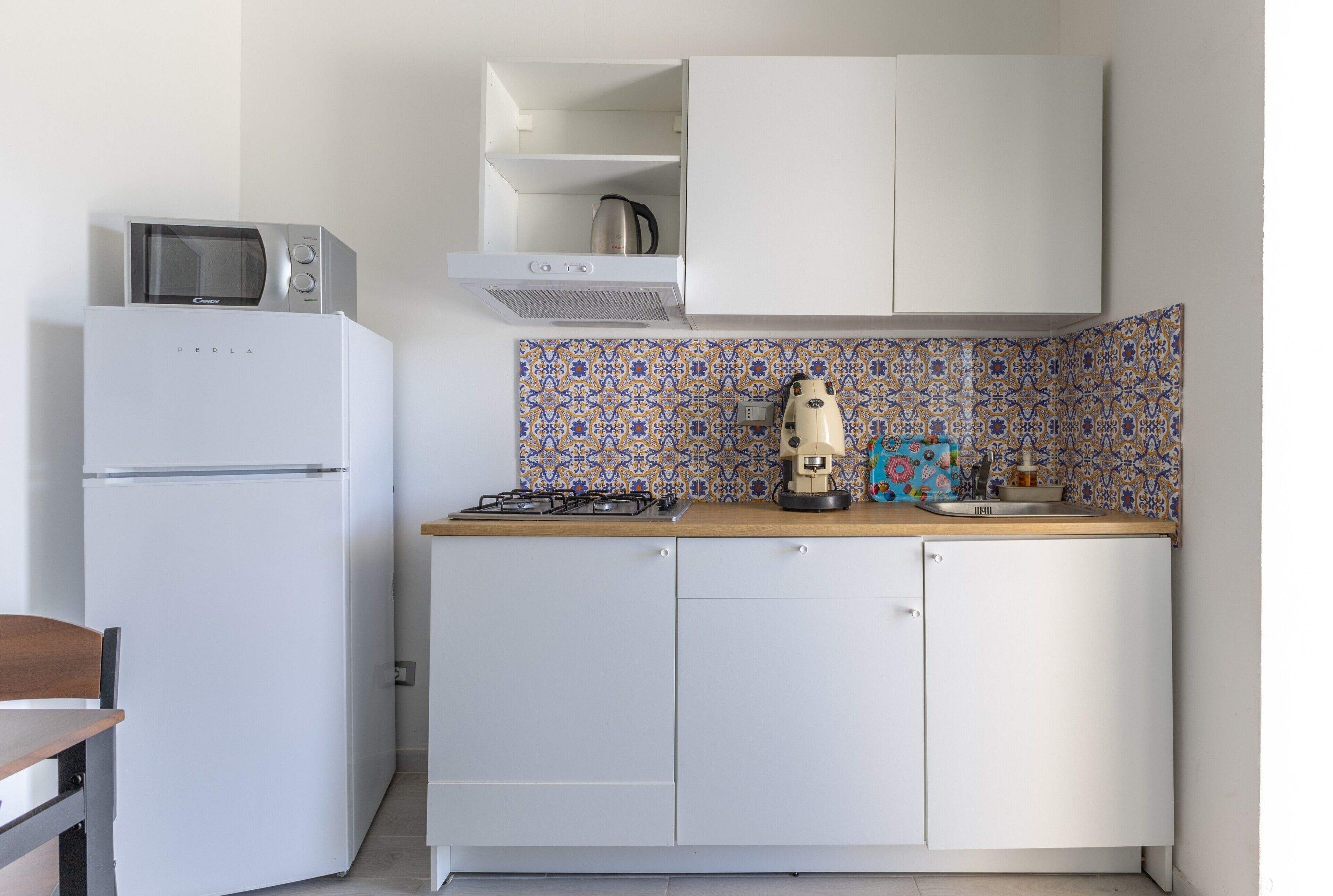 Een kleine keuken met een witte koelkast, een magnetron, een gasfornuis en een koffiezetapparaat.