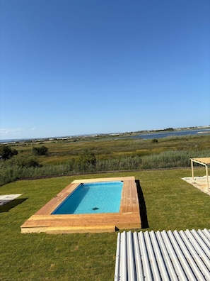 Ein rechteckiger Pool mit Holzterrasse, umgeben von Rasenflächen und Blick auf ein Gewässer.