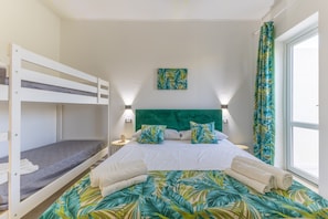 Ein Zimmer mit Etagenbetten, einem grünen, gepolsterten Kopfteil, einer Bettdecke mit tropischen Blättern und einem Fenster mit Vorhängen.