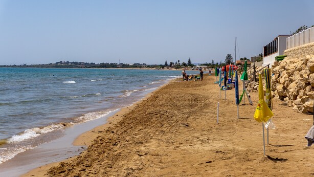 A sandy beach with a clear sky, a rocky wall, and a few umbrellas.