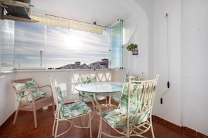 Un coin repas avec une table en verre et des chaises en osier, une fenêtre donnant sur des immeubles et une plante en pot accrochée au mur.