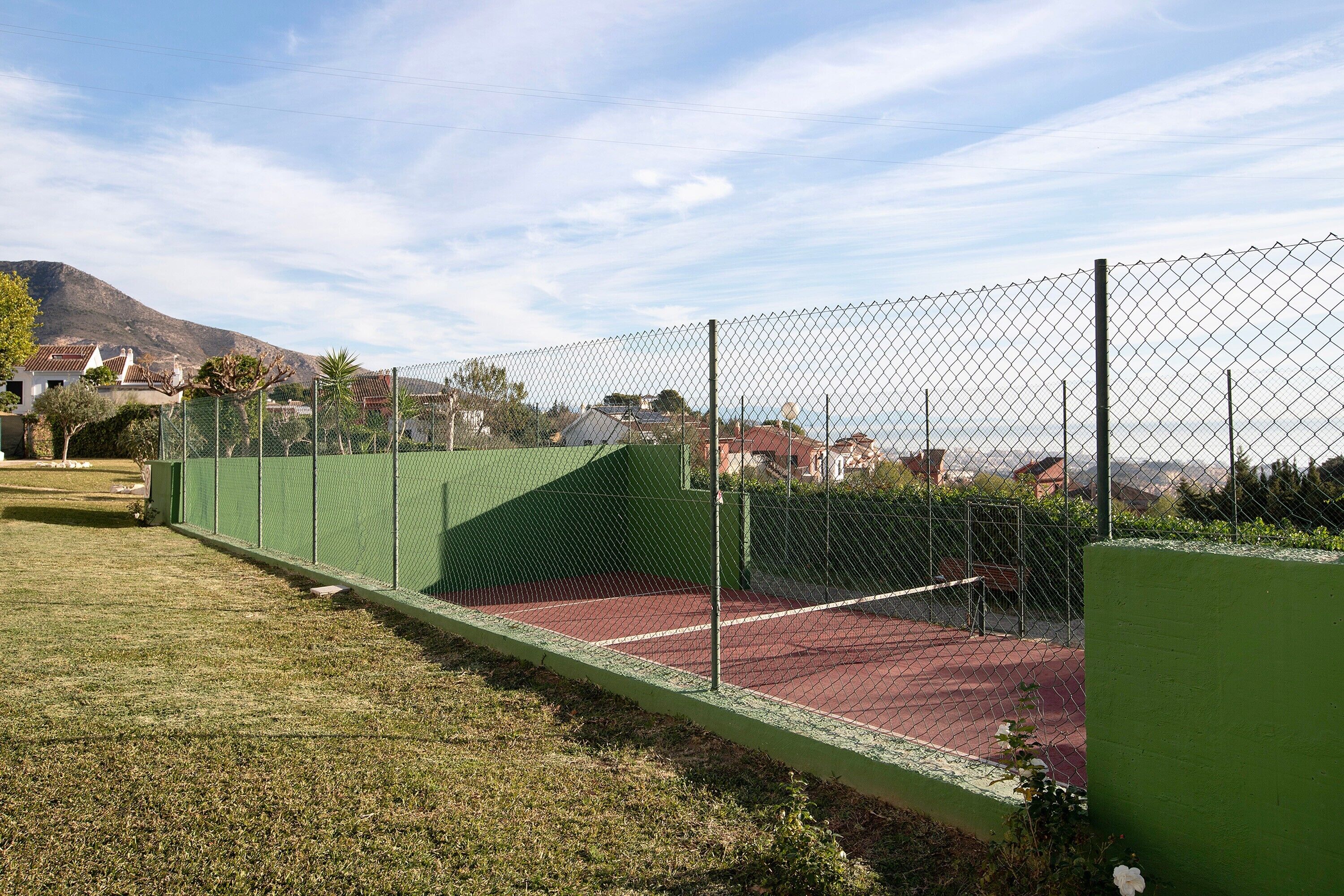 Una cancha de tenis con cerca verde, superficie roja y montañas al fondo.