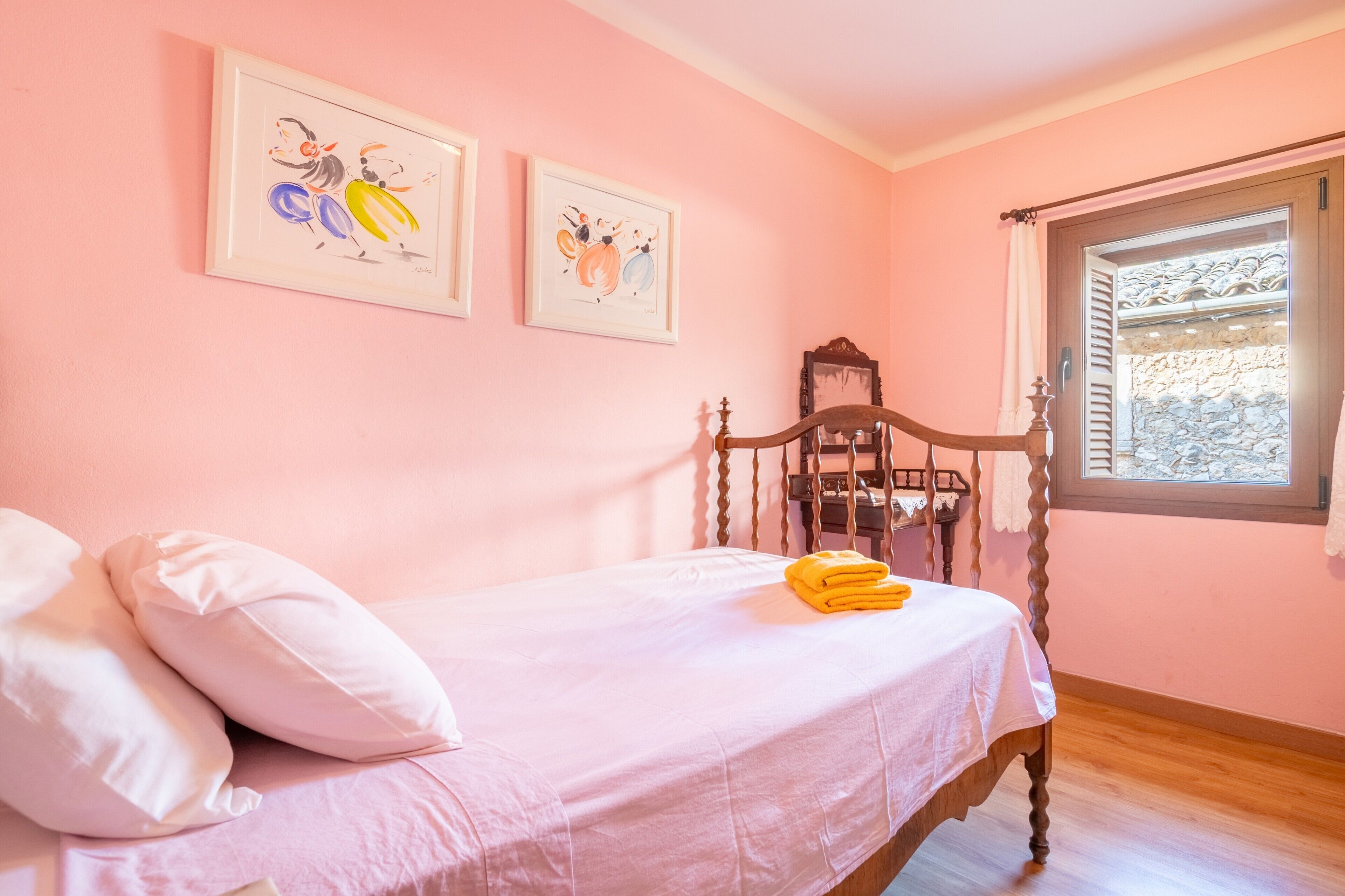 Un dormitorio con cama, almohadas, una silla de madera, una ventana y dos obras de arte colgadas en la pared.
