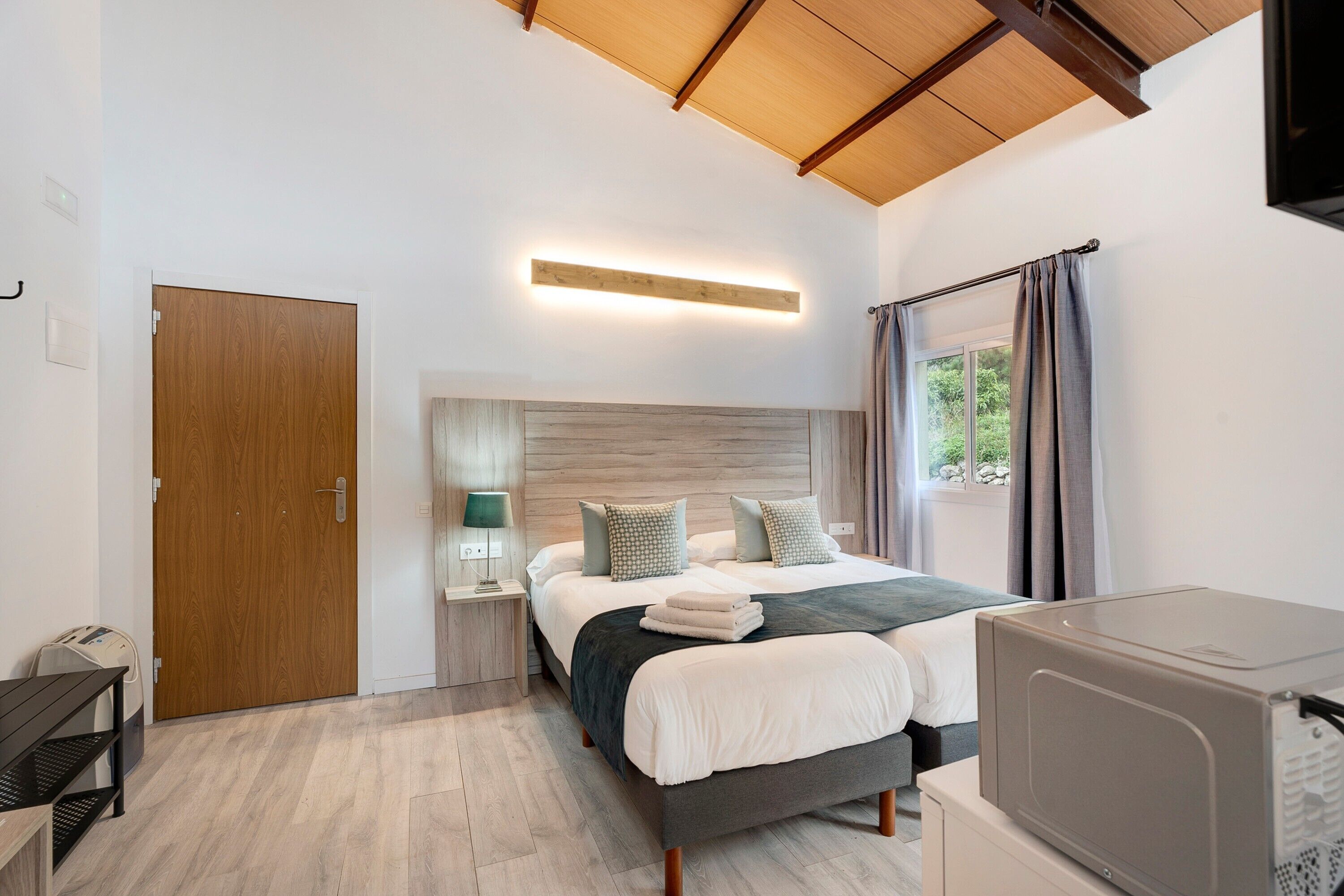 Une chambre à coucher avec un lit, des tables de chevet, un téléviseur, une porte en bois et une fenêtre avec des rideaux.