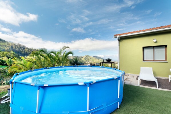 Une piscine recouverte d’une bâche bleue, une terrasse avec une chaise blanche et une maison avec un mur vert et une fenêtre.