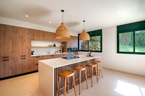 A modern kitchen with wooden cabinets, a white island with stools, pendant lights, and a view of greenery through a window. - Villa 'Bianca Tamariu' with Private Pool, Private Terrace and Wi-Fi (Tamariu)