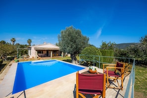 A swimming pool with a house in the background, surrounded by greenery and a clear sky.