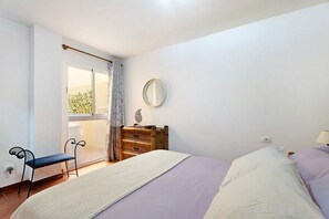 Ein Schlafzimmer mit Bett, Stuhl, Kommode, Spiegel und einem Fenster mit Vorhängen.
