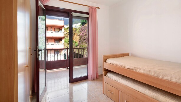 Un dormitorio con una cama litera de madera, un ventanal corredizo que da a un balcón y vistas de un edificio con balcones.