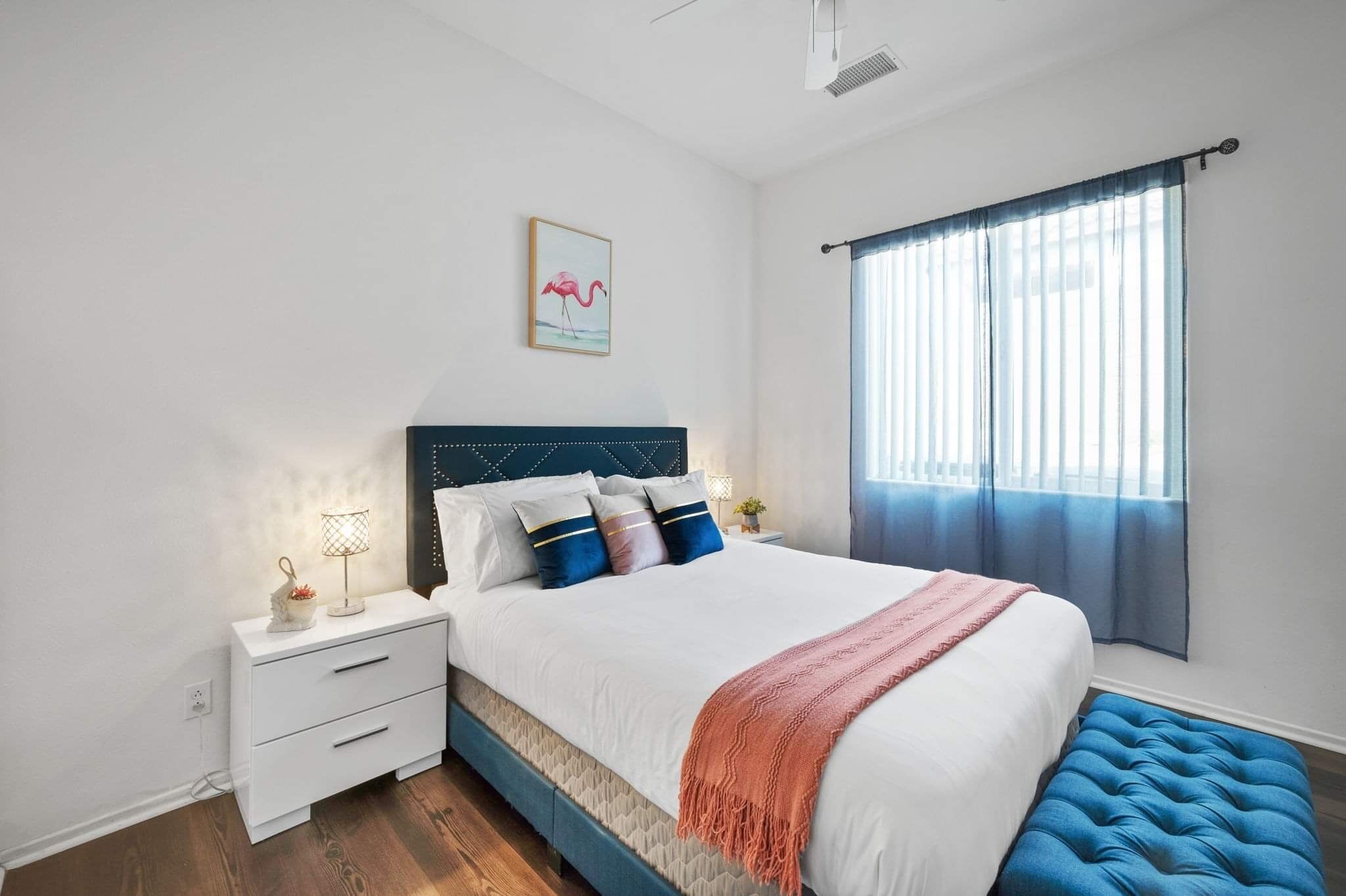 Una camera da letto con un letto, comodini, un tavolino di fianco al letto, una finestra con tende e un quadro appeso al muro.