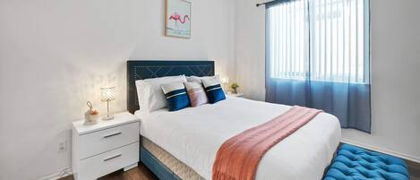Una camera da letto con un letto, comodini, un tavolino di fianco al letto, una finestra con tende e un quadro appeso al muro.