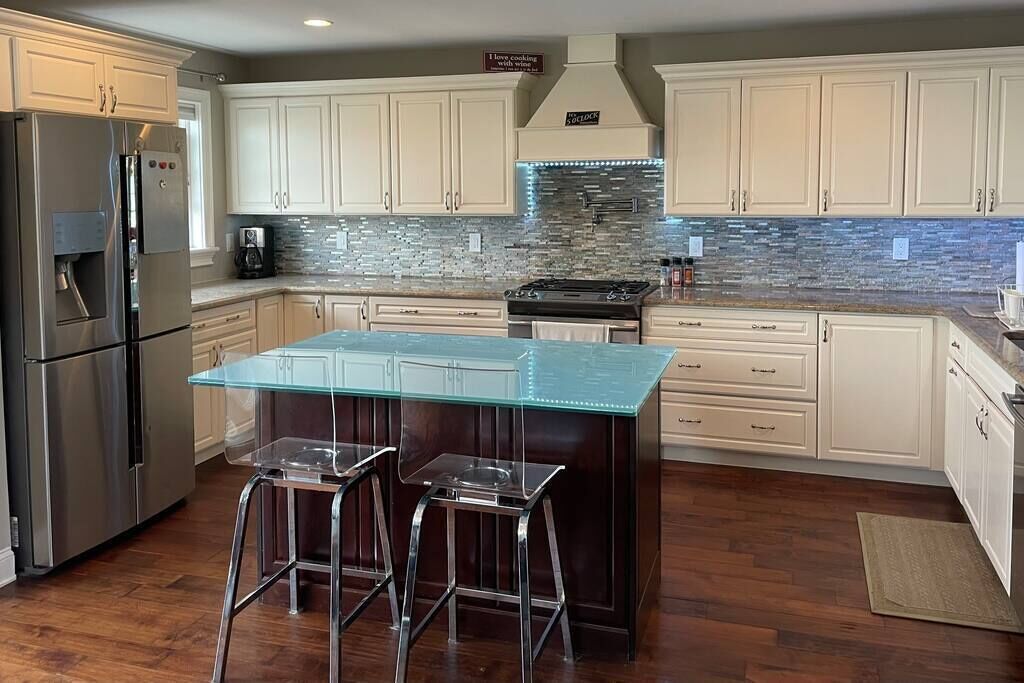 A modern kitchen with white cabinets, a stainless steel refrigerator, a glass island with stools, and a tiled backsplash.