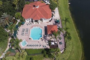 An aerial view of a resort with a large swimming pool, multiple lounge areas, and a red-roofed building.