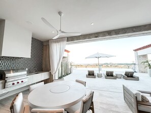 Een moderne keuken met een inbouwfornuis, een eethoek met een ronde tafel en stoelen, en een terras met buitenmeubilair en uitzicht op het landschap.