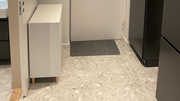Ein kompakter Raum mit einer weißen Tür, einem schwarzen Kühlschrank, einem kleinen weißen Schrank und einer grauen Matte auf dem Boden.