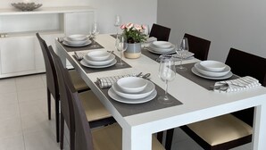 Ein moderner Essbereich mit einem weißen Tisch, gedeckt für sechs Personen, inklusive Teller, Schüsseln und Besteck. Dunkle Holzstühle und ein Blumenstrauß in einer Vase stehen auf dem Tisch.