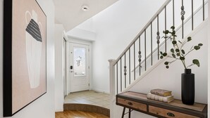A hallway with a wooden floor, a staircase with a white railing, a wall-mounted artwork, and a console table with books and a vase. - Cozy Home w/ Free Parking | Fireplace & Backyard | Sleeps 8 | Near Banff (Calgary)