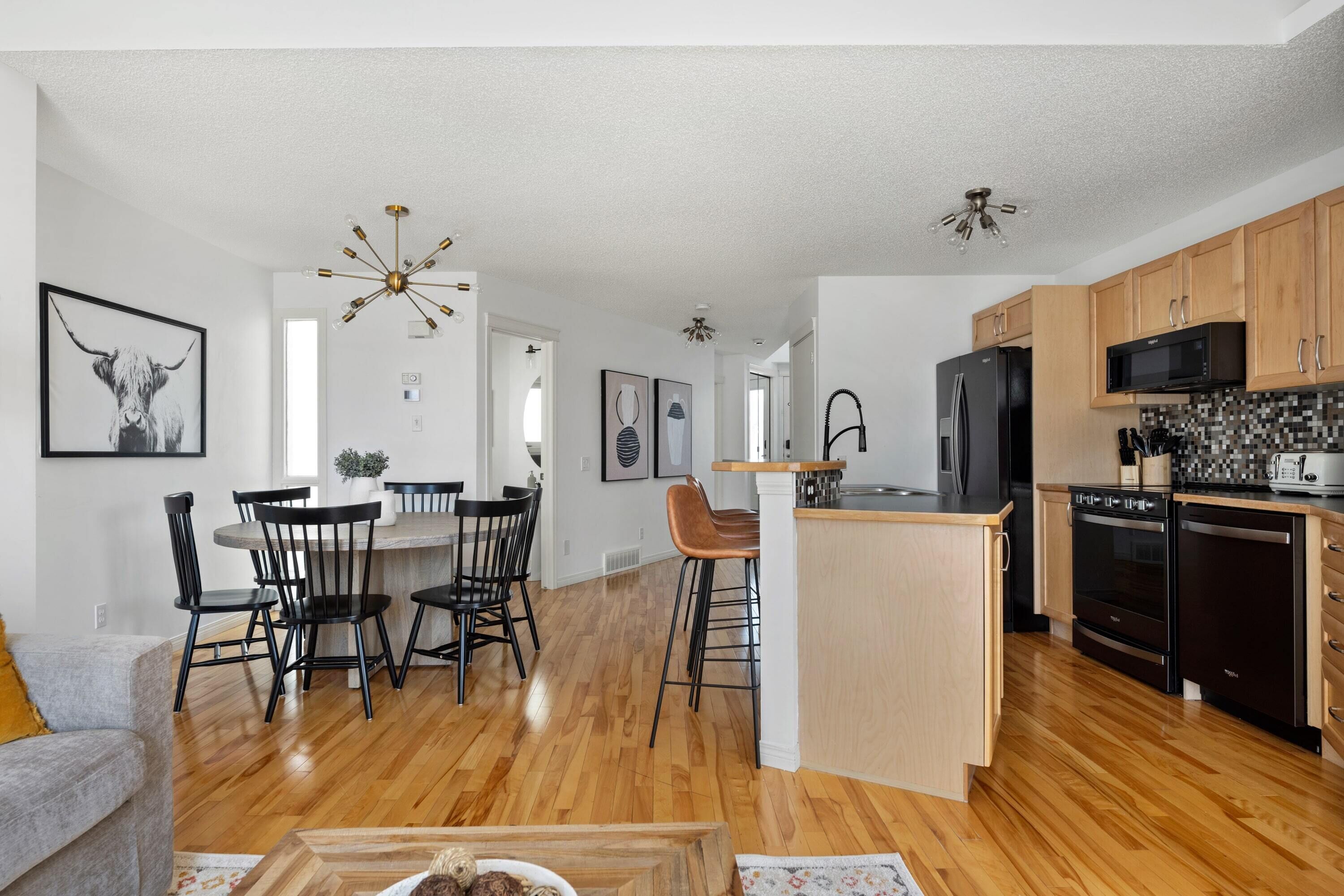 A modern kitchen with wooden cabinets, a black refrigerator, and a microwave. There is a dining area with black chairs and a wooden table.