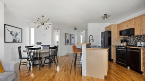 A modern kitchen with wooden cabinets, a black refrigerator, and a microwave. There is a dining area with black chairs and a wooden table. - Cozy Home w/ Free Parking | Fireplace & Backyard | Sleeps 8 | Near Banff (Calgary)