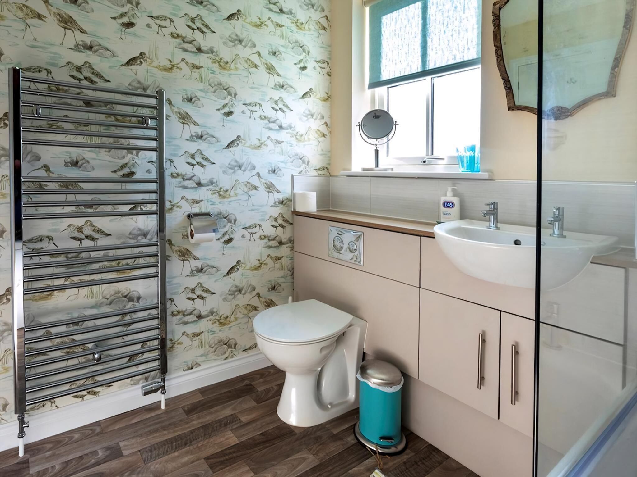 A bathroom with a toilet, sink, and mirror. There is a towel rack and a trash bin.