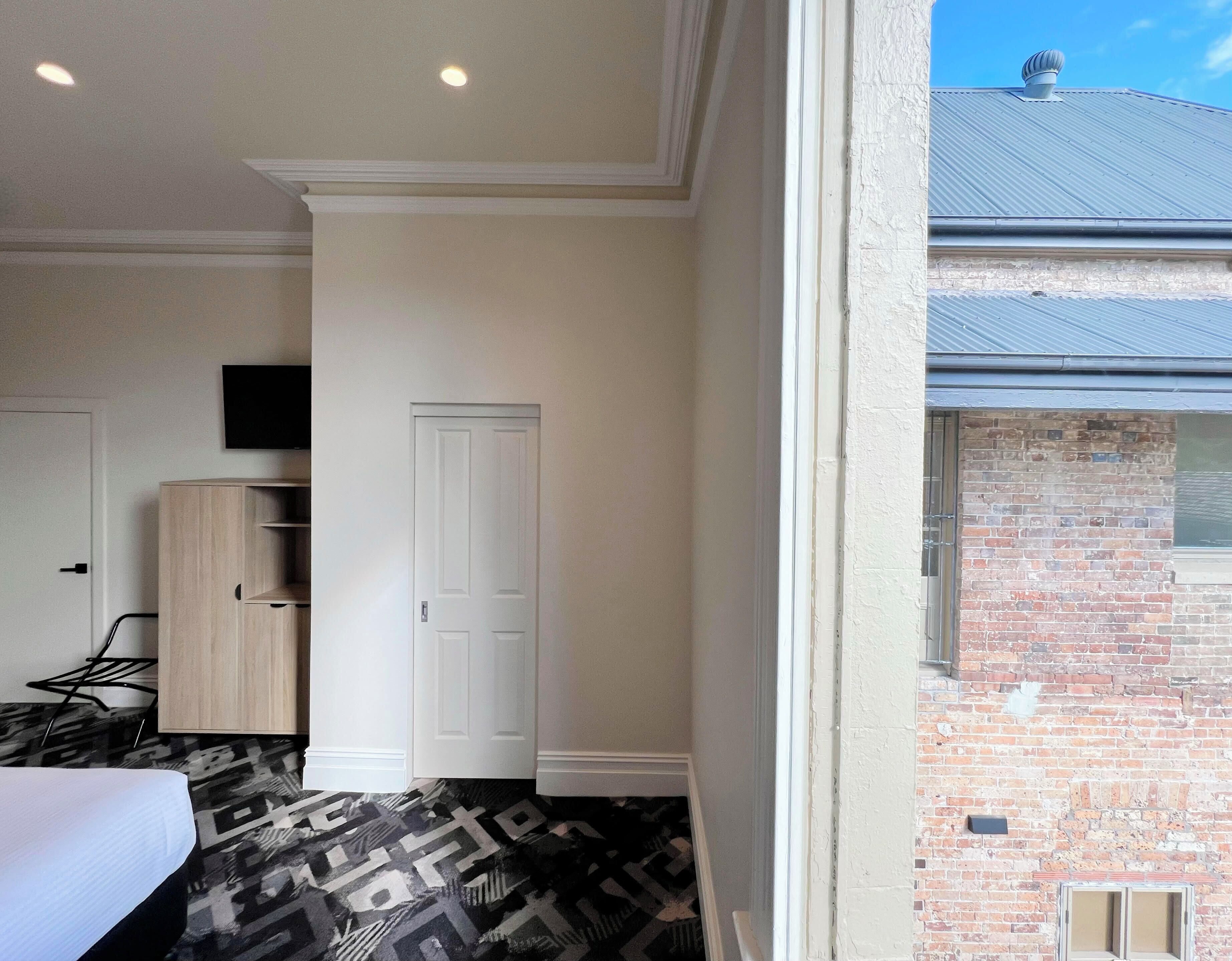A hotel room with a bed, a wardrobe, a television, and a brick wall visible through the window.