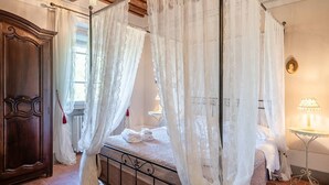 A four-poster canopy bed with white linens, a wooden wardrobe, and a small table with a lamp.