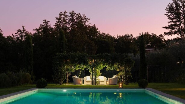 A swimming pool with two lounge chairs and a small table, surrounded by trees and a building in the background.