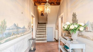 A hallway with a staircase, a chandelier, a vase of flowers, and a mural on the walls.