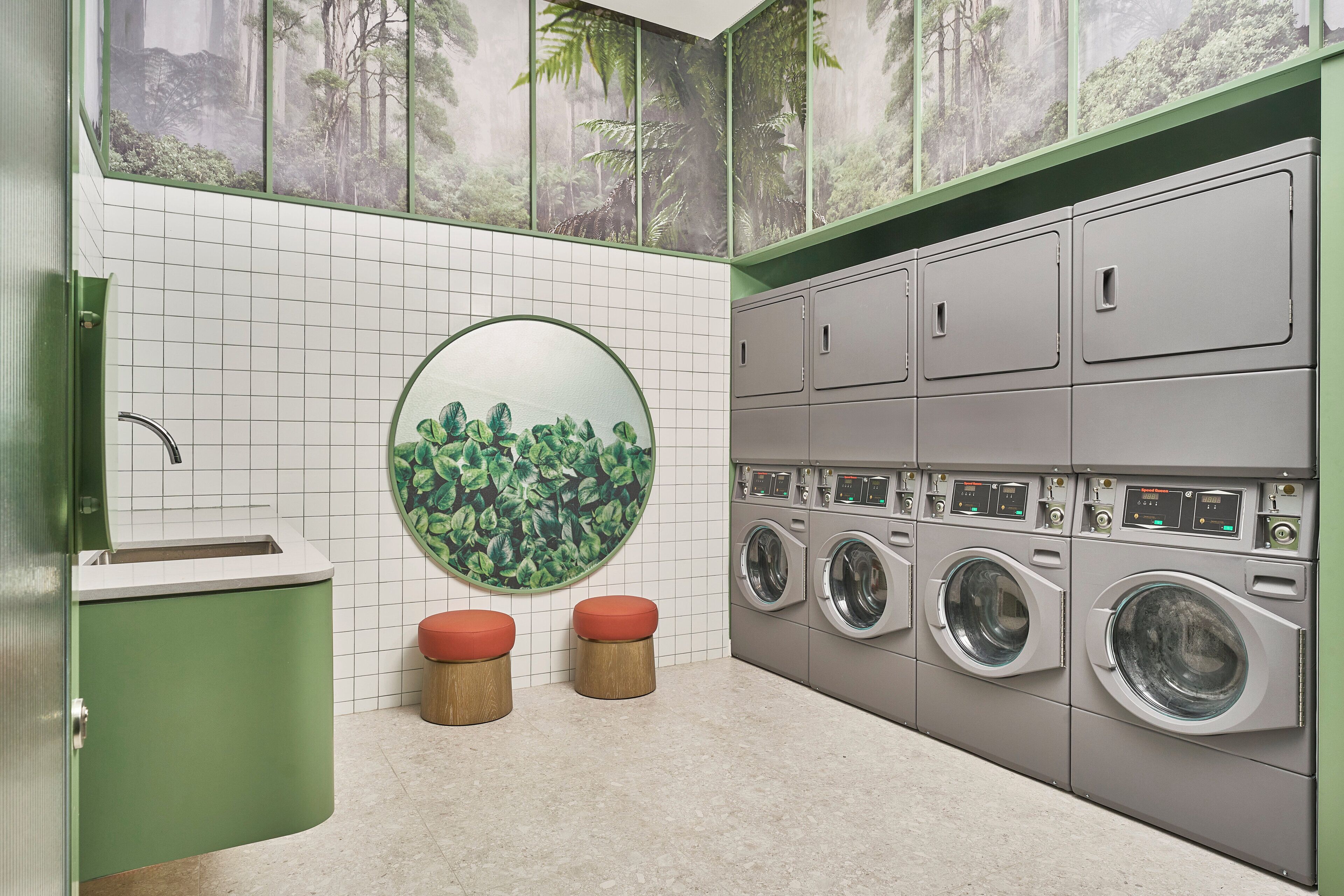 Una sala de lavandería con un lavamanos verde, dos taburetes, un espejo circular grande y varias lavadoras.