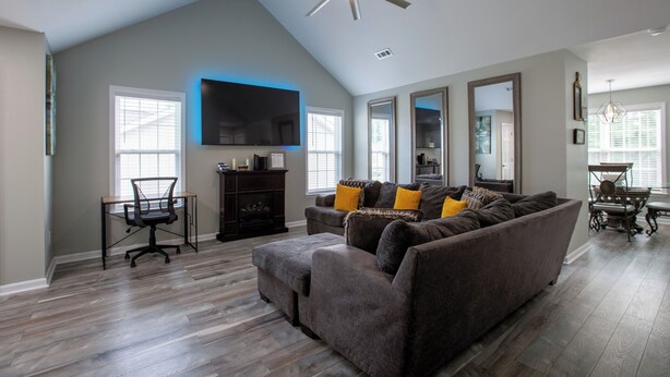 A spacious living room with a ceiling fan, a large flat-screen TV, a dark brown sofa with yellow cushions, and a wooden dining table with chairs.