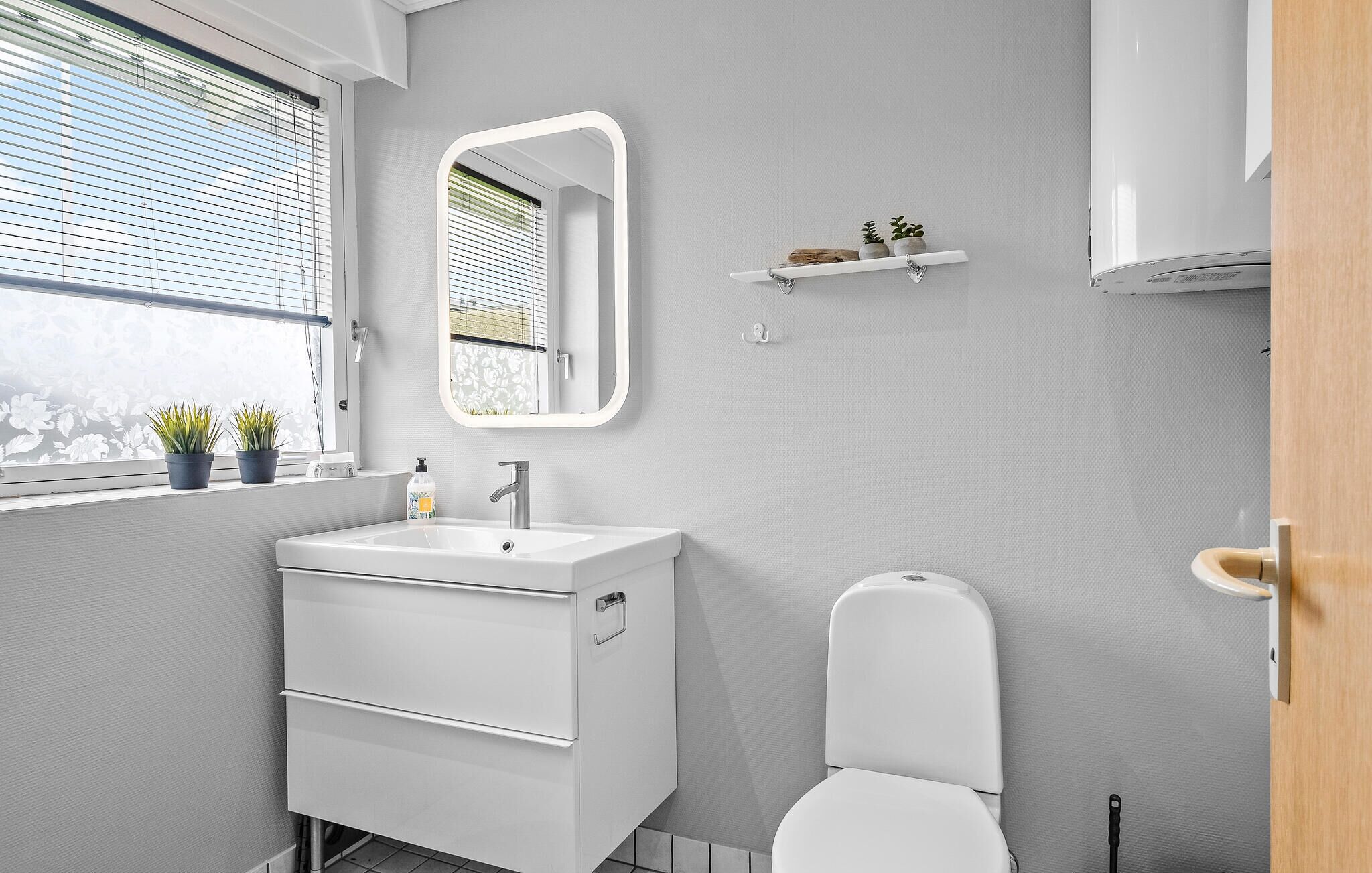A modern bathroom with a white sink, mirror, and toilet. There are potted plants on the windowsill and a small shelf with toiletries.