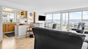 Una sala de estar moderna con un sofá gris, un comedor con mesa y sillas, una cocina con armarios de madera y un ventanal con vista al mar.