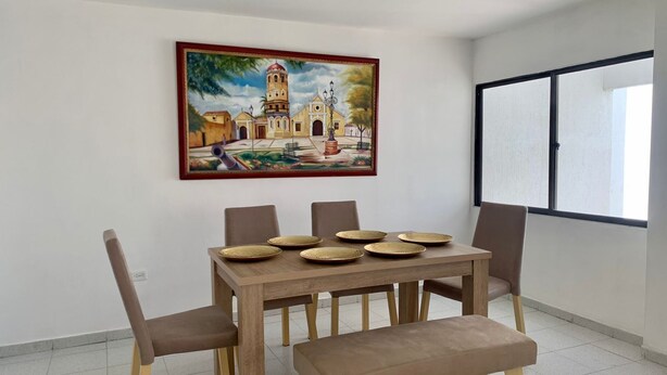 A dining area with a table set for four, surrounded by chairs and a bench. A painting of a historical building is on the wall. - «Javi's house: explore the charm of Cartagena (Cartagena de Indias)
