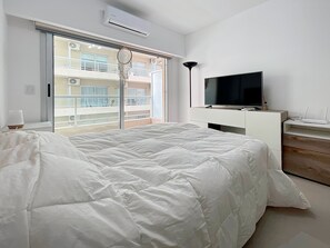 A bedroom with a large bed, a TV on a white dresser, and a balcony with a view of other buildings.