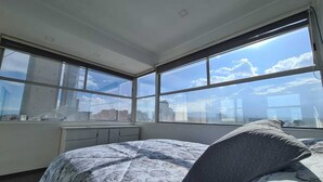 Ein modernes Schlafzimmer mit einem großen Bett und Blick auf die Stadt durch raumhohe Fenster. Draußen ist eine Stadtansicht zu sehen.