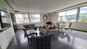 Ein modernes Wohnzimmer mit einer Couch, Sesseln, einem Couchtisch und einem Fernseher. Große Fenster bieten Blick auf die Stadt.