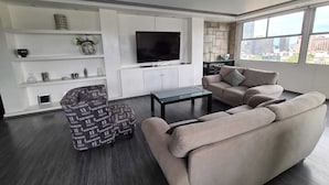 Ein modernes Wohnzimmer mit Sofa, Hocker und Couchtisch. Ein Flachbildfernseher ist an der Wand montiert, und ein Fenster bietet Blick auf die Stadt.