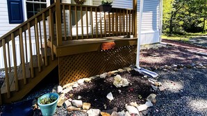 Un porche cubierto con baranda de madera, un pequeño jardín con rocas y una maceta, y un edificio blanco con contraventanas azules.