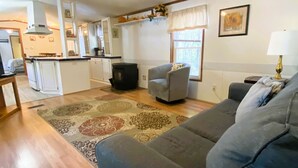 Una sala de estar con un sofá, una silla, un tapete, un ventilador de techo y una cocina al fondo.