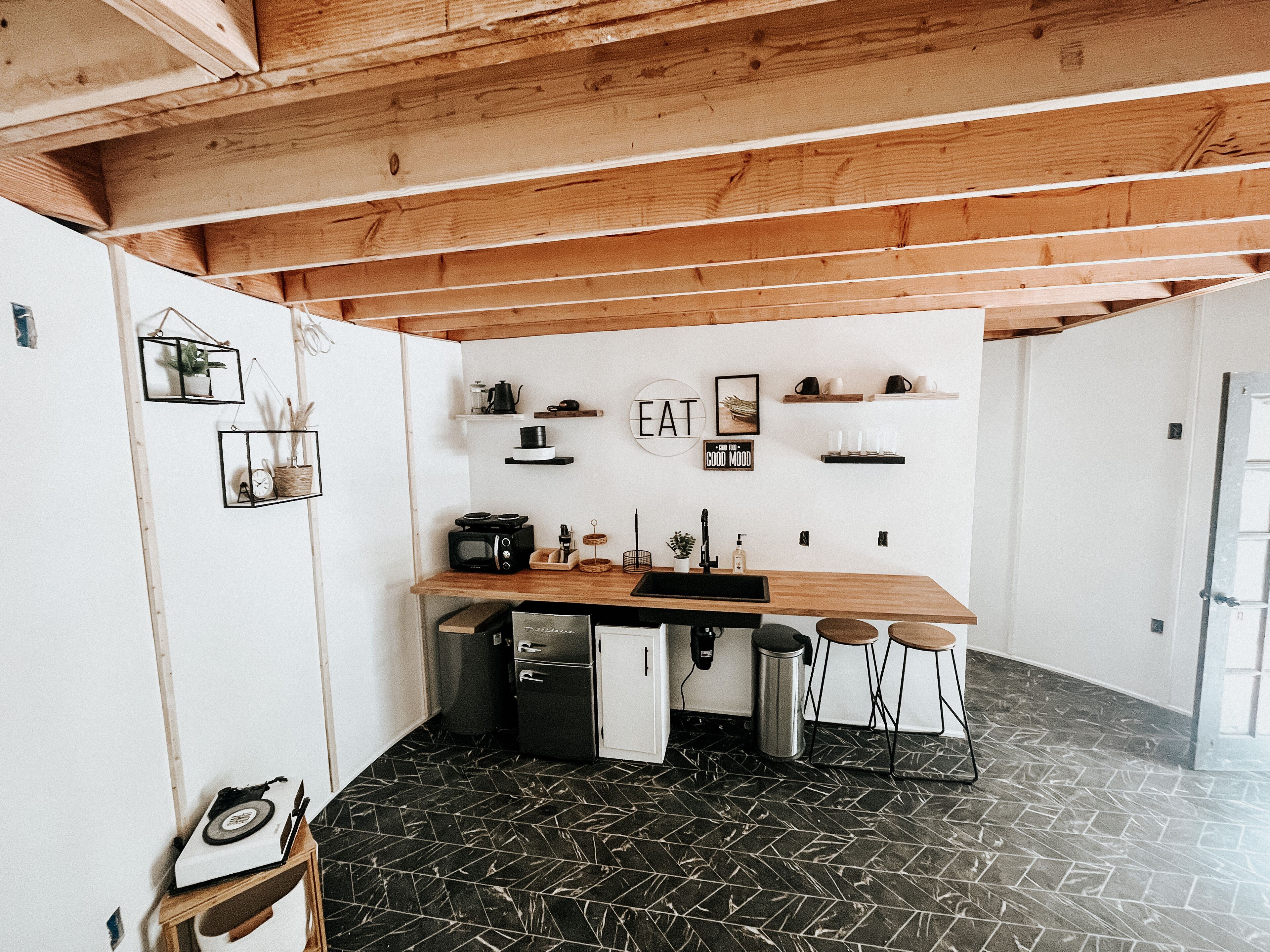 Une cuisine compacte avec un plan de travail en bois, un évier et un four à micro-ondes. On y trouve deux tabourets et un petit réfrigérateur. Le sol est recouvert d’un motif géométrique.