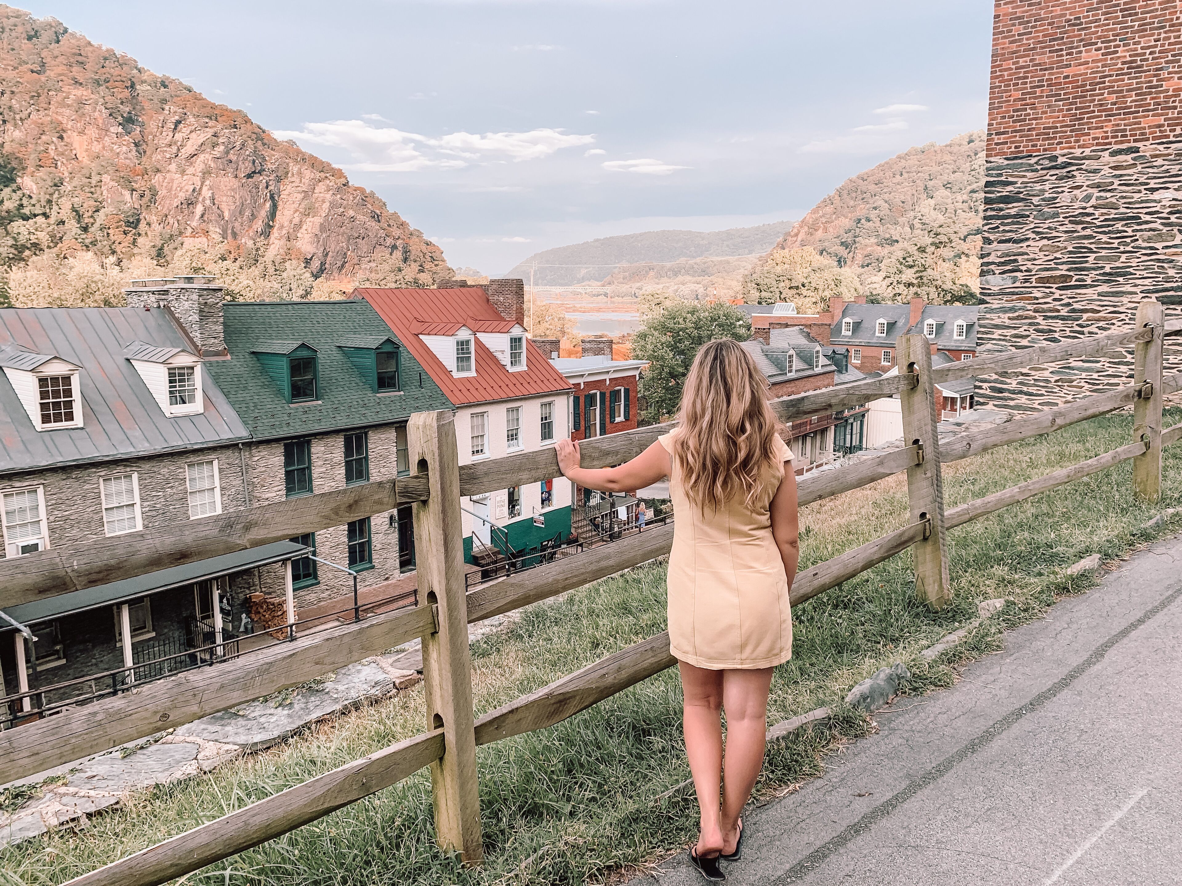Eine Frau steht auf einer Holzbalustrade und blickt auf eine Stadt mit historischen Gebäuden und einem Bergpanorama.